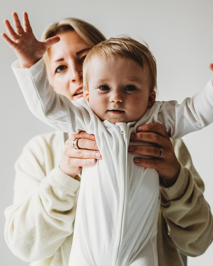 Cool mum holds beautiful baby girl, wearing plain white onesie with zip and with her arms outstretched
