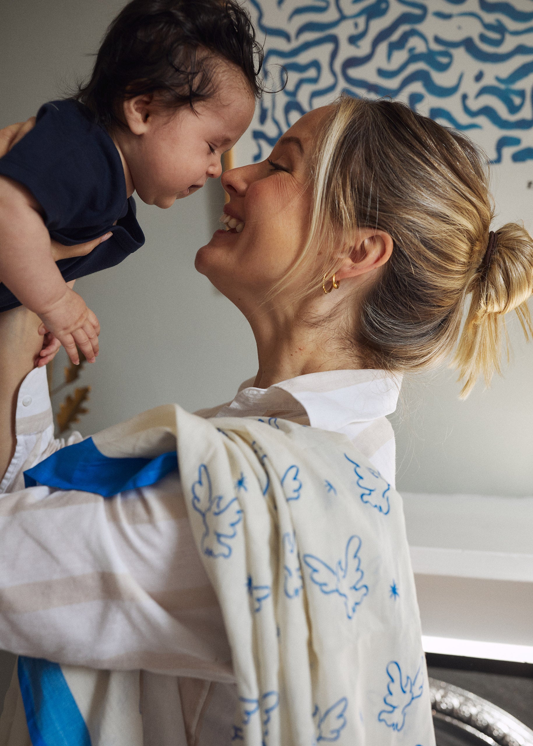 Stylish mum holds her baby girl in the air and kisses her nose, with an organic animal print muslin over her shoulder.