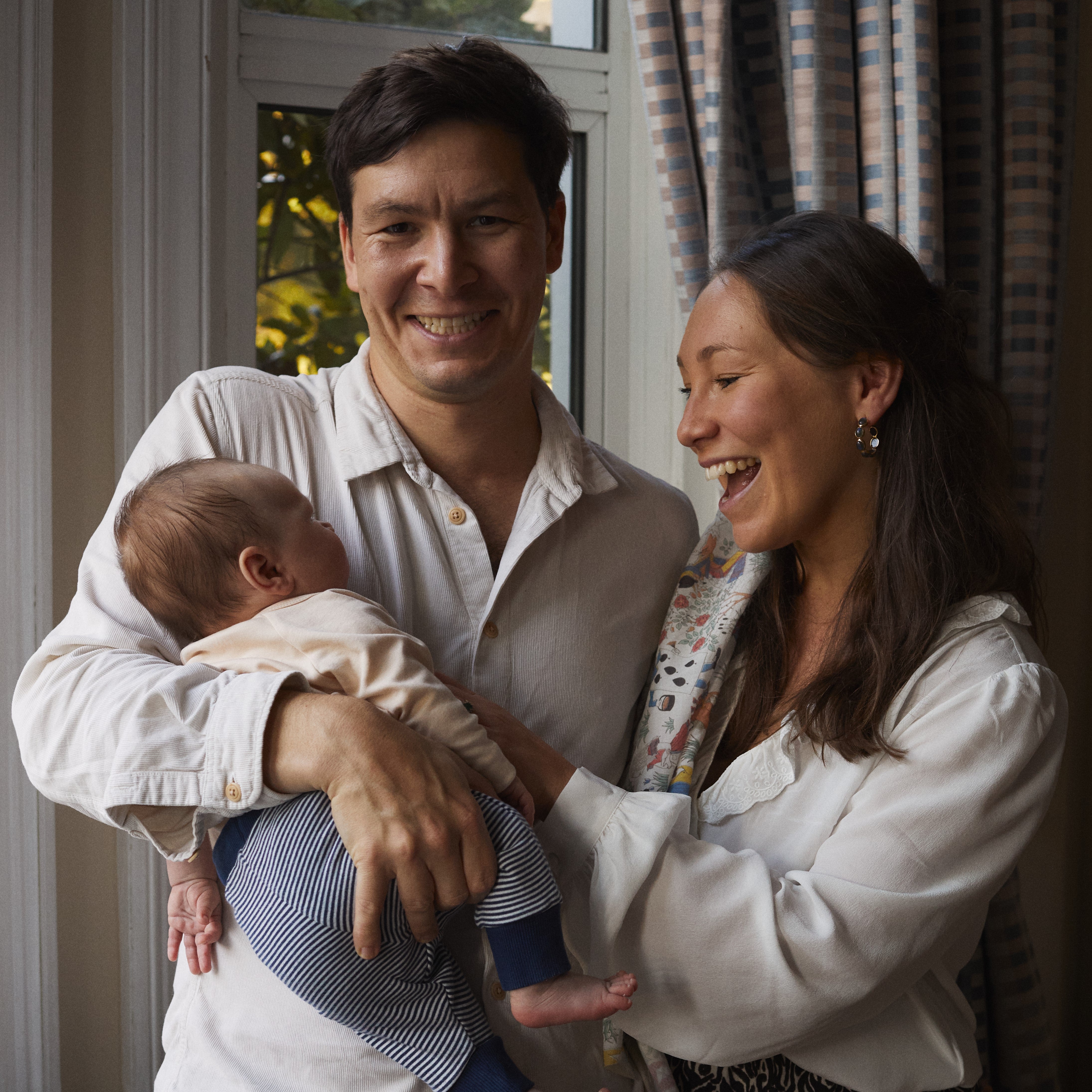 NINA founders Emily & Oli with their newborn baby boy standing together for a family photo with a NINA muslin over Emily’s shoulder