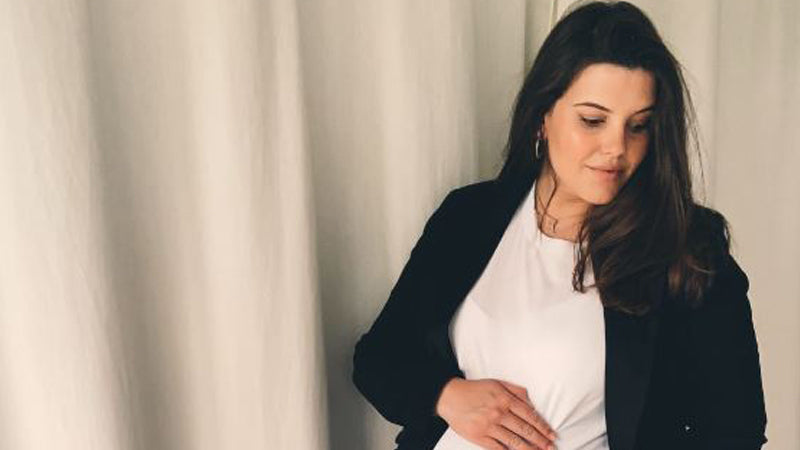 Pregnant woman stands in front of curtains holding her bump on her due date..